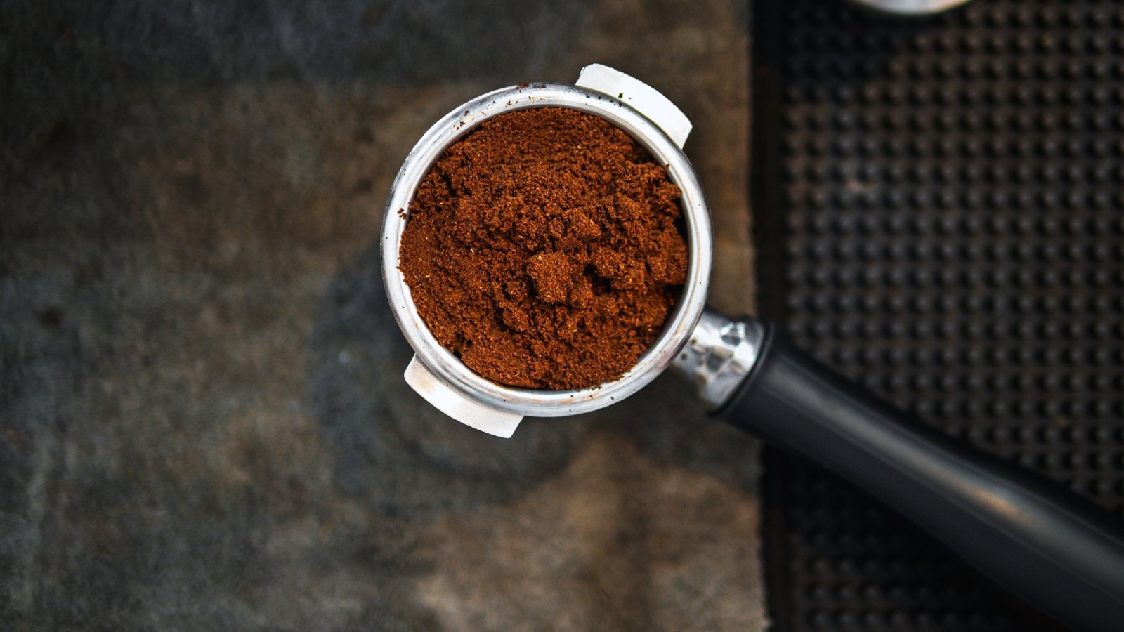 A portafilter filled with fine, compacted compost placed on a dark, textured surface.