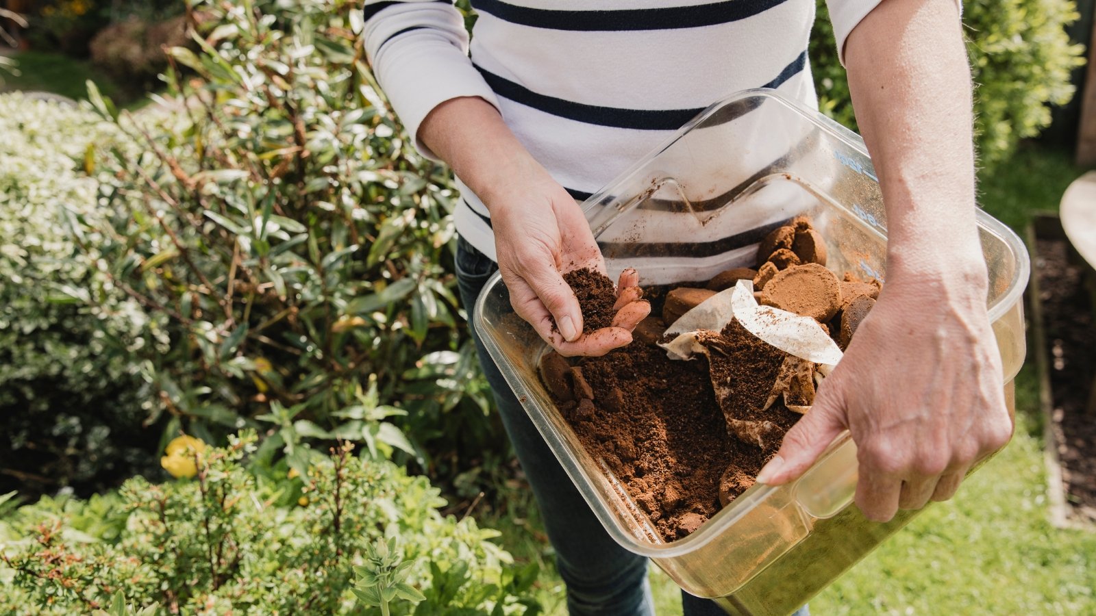 A person wearing a striped shirt mixing organic compost in a container outdoors, surrounded by green foliage.