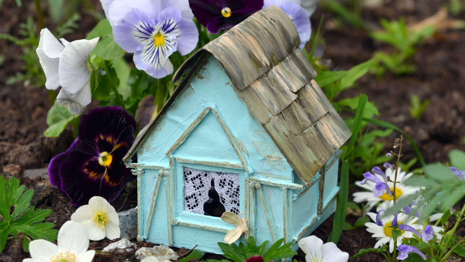 Viola x wittrockiana 'Cool Wave' with purple and white flowers near a small blue house model with a thatched roof.