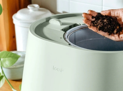 A hand holds dark, processed organic waste over a modern kitchen decomposer on a countertop, looking like the Lomi composter