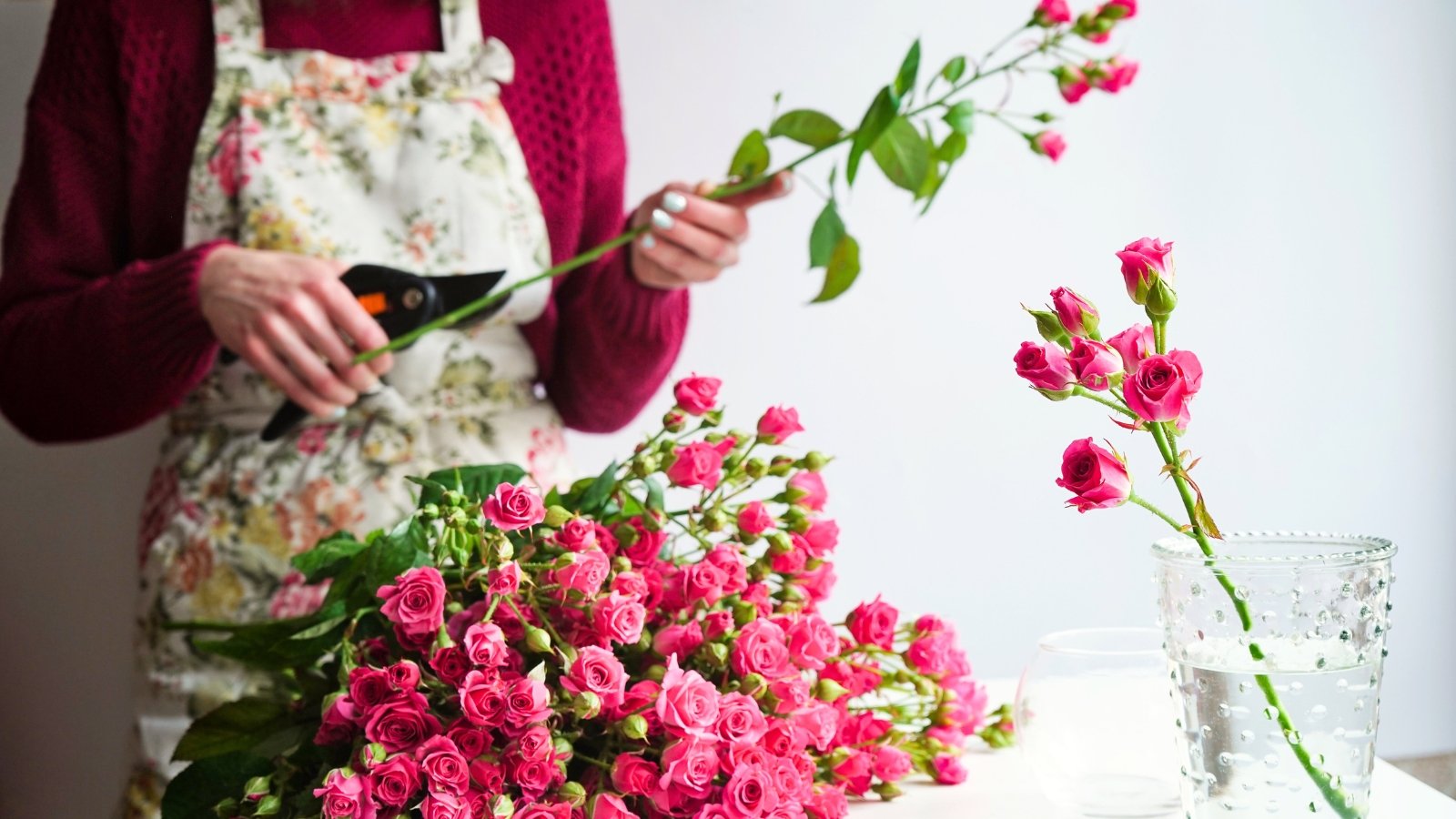 A woman in a colorful apron trims the lower leaves and stem tips of a bush rose with clusters of small pink flowers, placing them in a vase of clean water next to a large bunch of freshly cut flowers.
