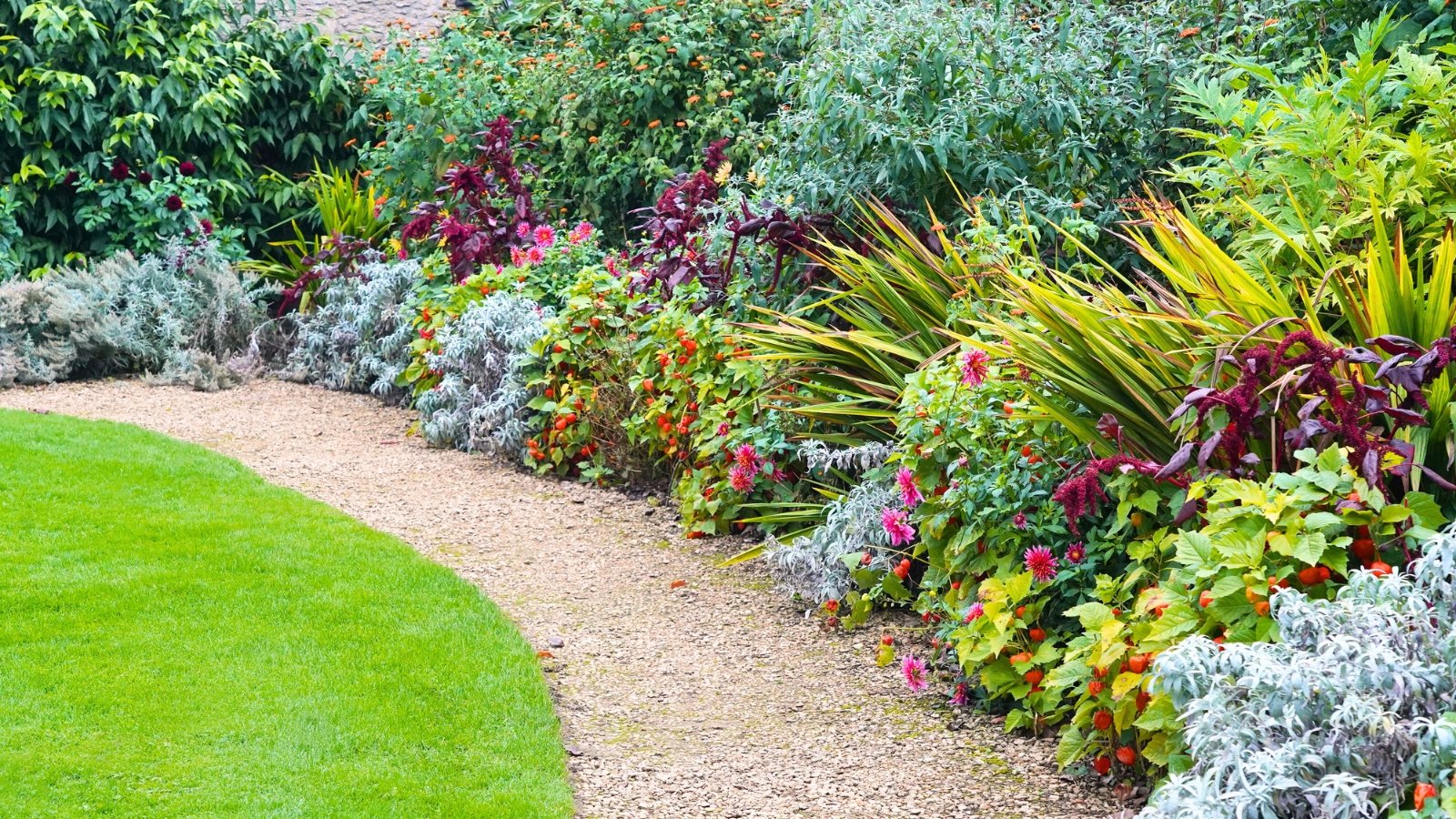 A beautiful garden with a colorfully blooming flowerbed separated by a winding path that turns into a green lawn.