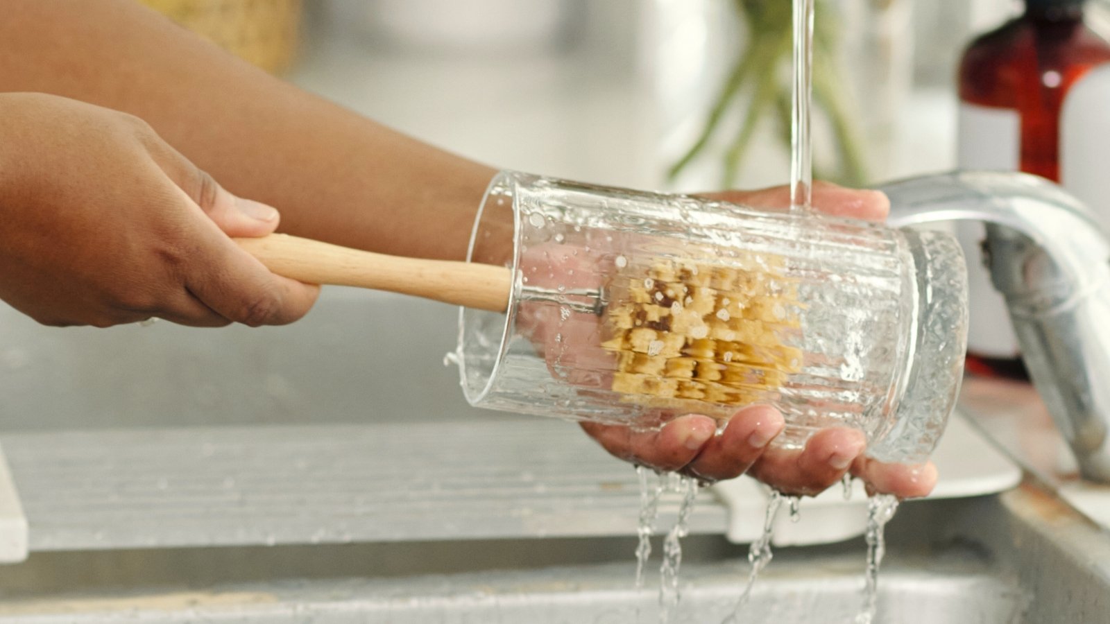 A woman washes a glass vase with an eco-brush in a sink in a bright kitchen.