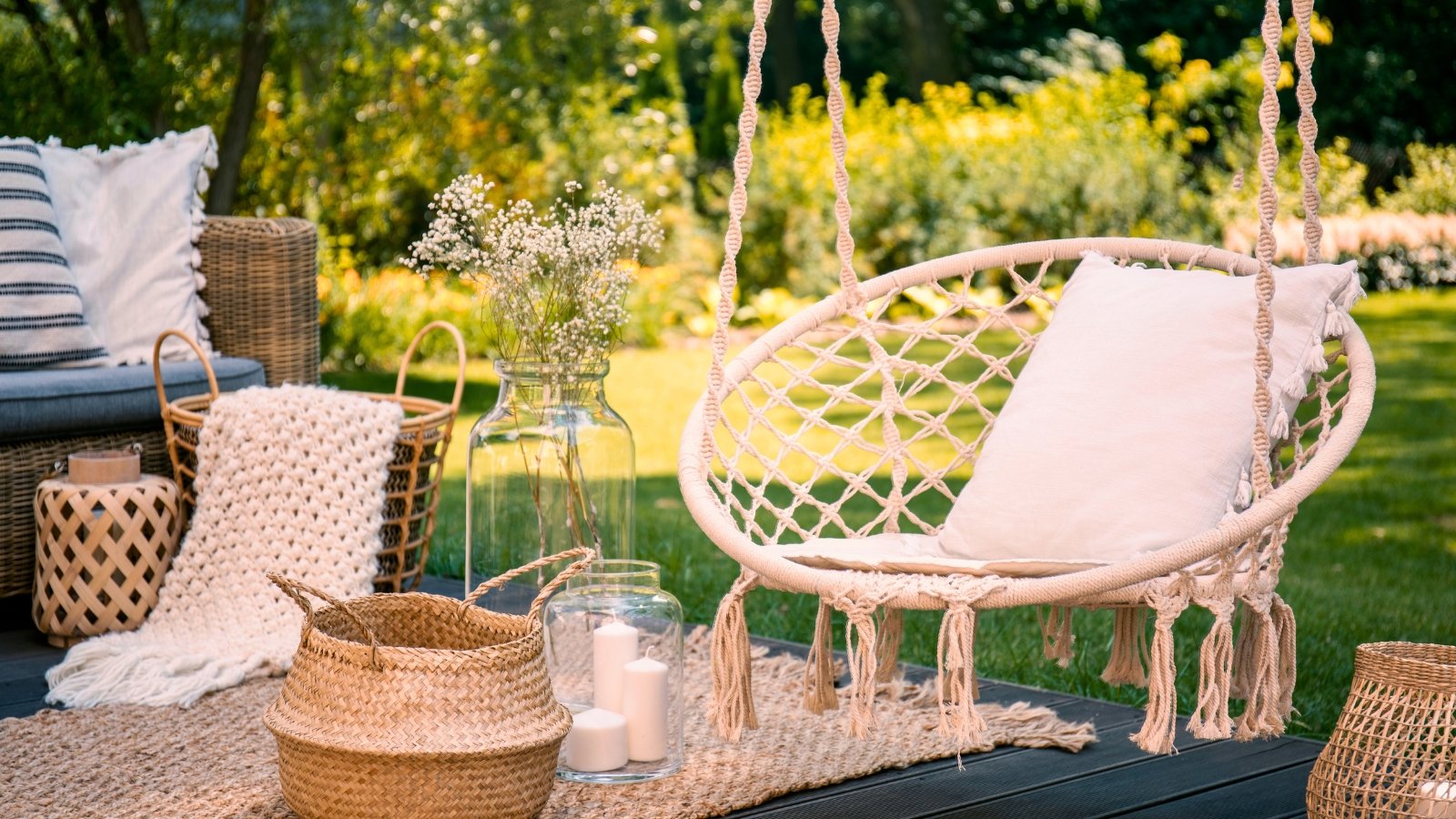 Garden seating area with sofa, hanging chair, rug and various rustic decor, surrounded by green lawn and various shrubs and trees.
