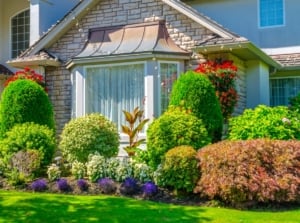 Stunning large classic style home with neat front garden including various evergreen, perennial and annual plants in flowerbeds and hanging containers surrounded by neat green lawn - demonstration of avoidance of basic landscape design mistakes.