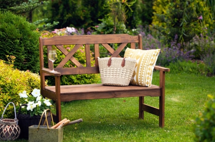 Upgraded backyard landscaping, rustic wooden bench with throw pillow, wicker bag and wine glass stands in the middle of green lawn surrounded by various bushes, trees and flowering plants