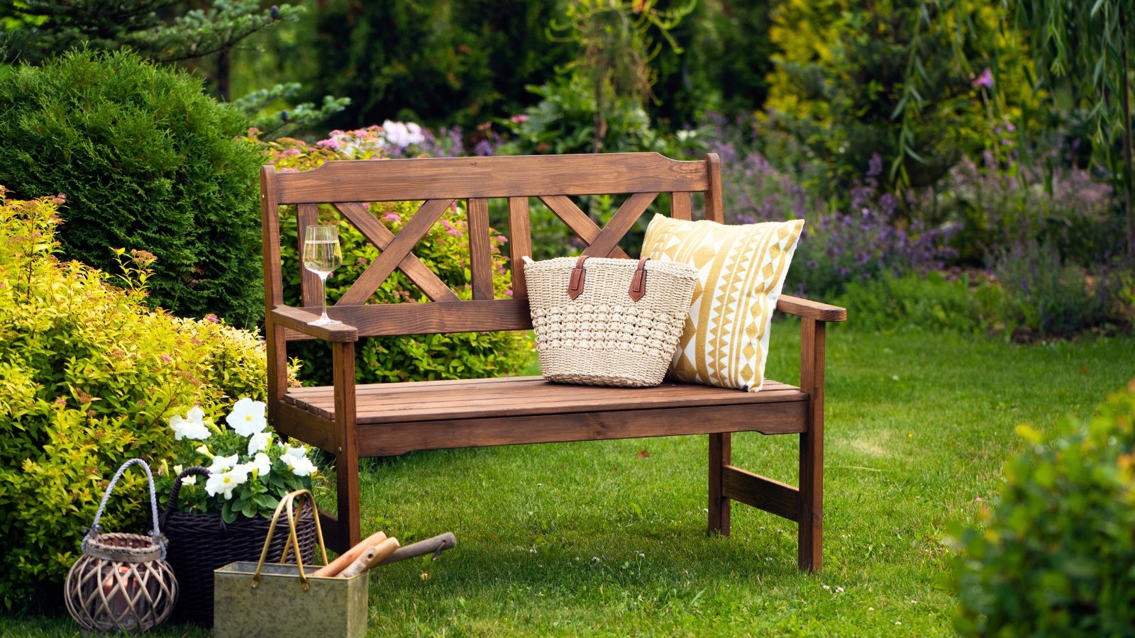 Upgraded backyard landscaping, rustic wooden bench with throw pillow, wicker bag and wine glass stands in the middle of green lawn surrounded by various bushes, trees and flowering plants