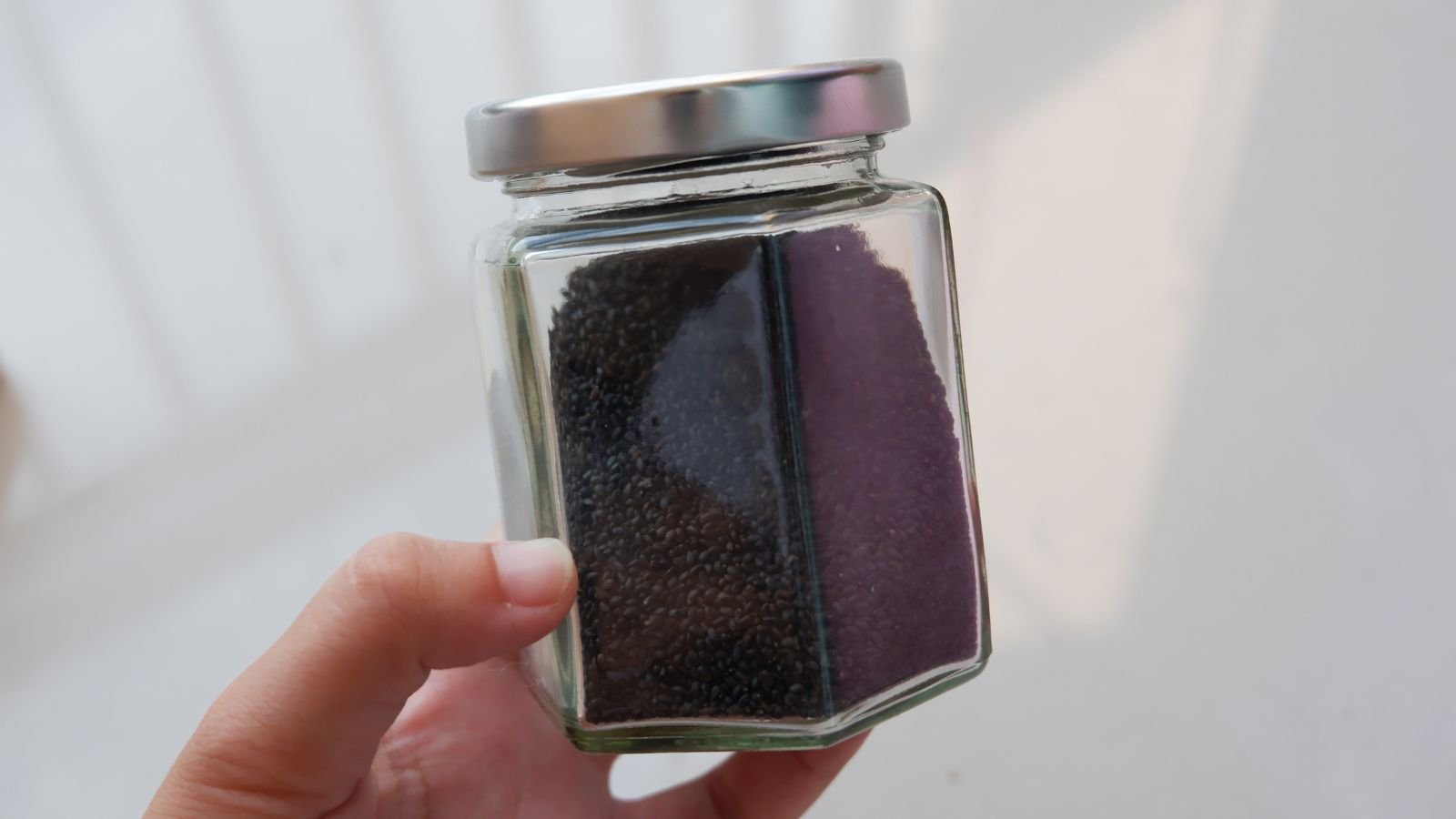 A close-up shot of a person's hand holding a glass container filled with small, black, ovules of an herb, in a well lti area indoors