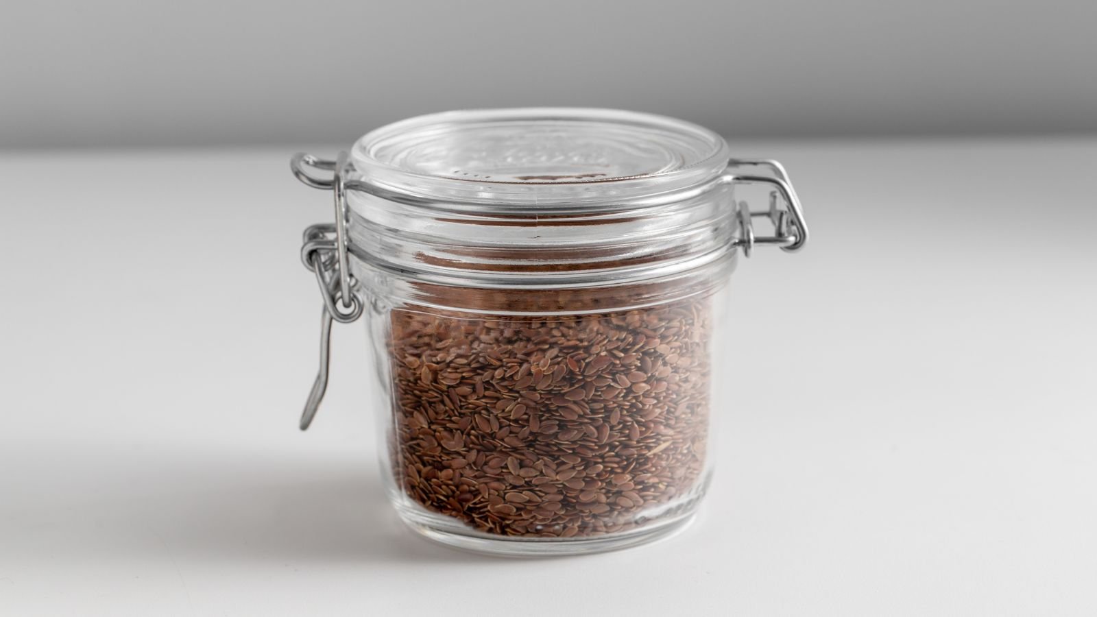 A close-up shot of an airtight jar filled with small brown colored seeds, placed on top of a white surface in a well lit area indoors