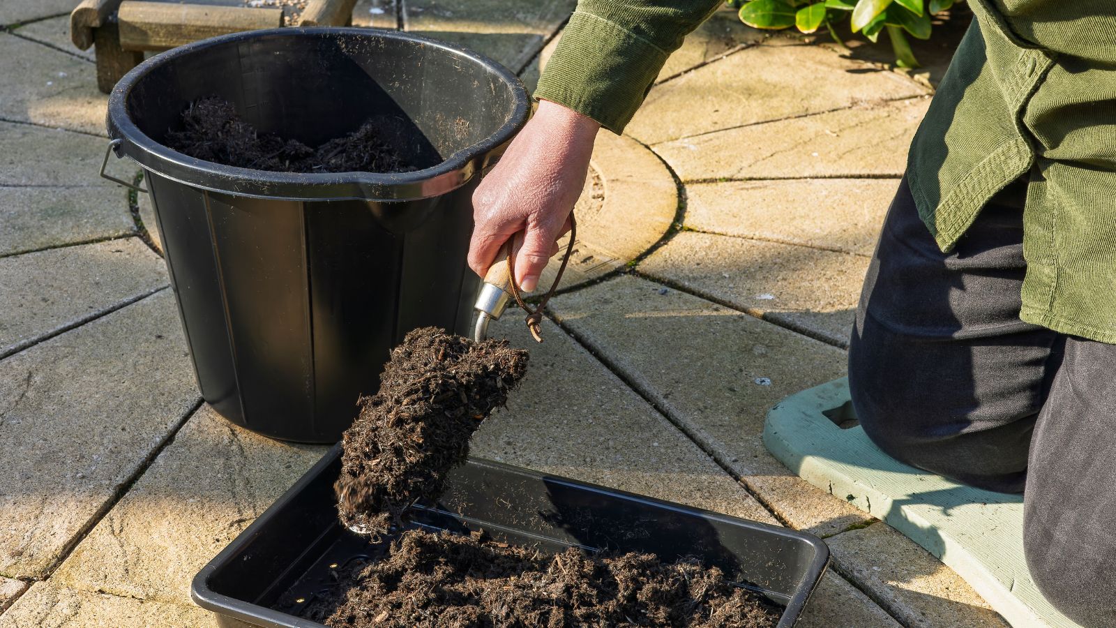 A gardener mixing soil kneeling beside a black bucket, placed in an area with sunlight