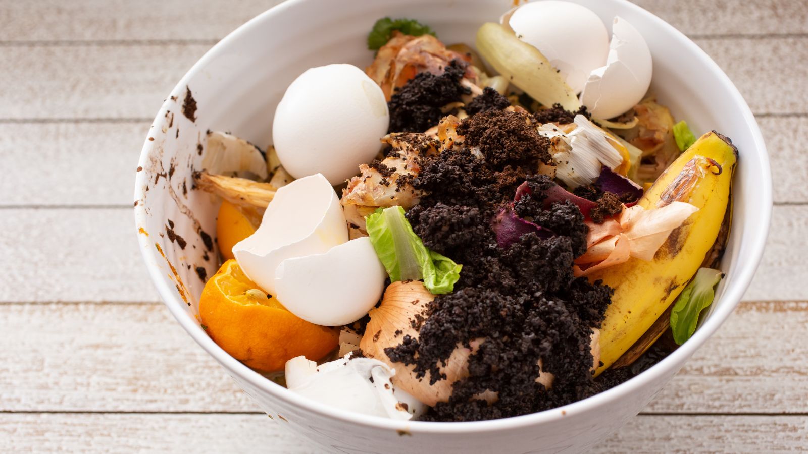 A shot of a bowl of compost, with various kitchen scraps and other sediments placed on tp of a wooden surface indoors