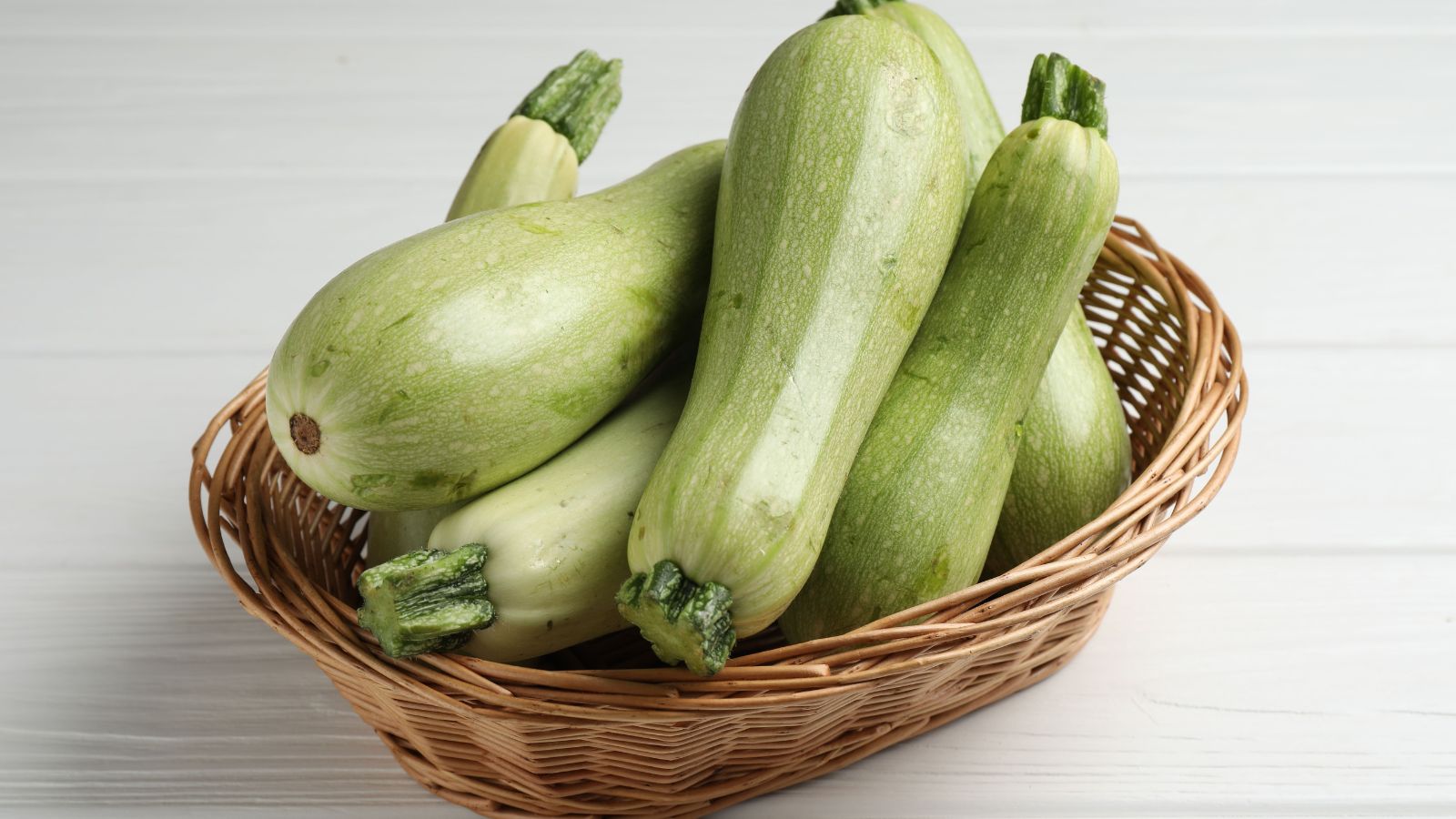 A close-up shot of a woven wicker basket holding a pile of white to pale-green colored fruits of the Cucurbita pepo ‘Bianca Goriziana’ variety