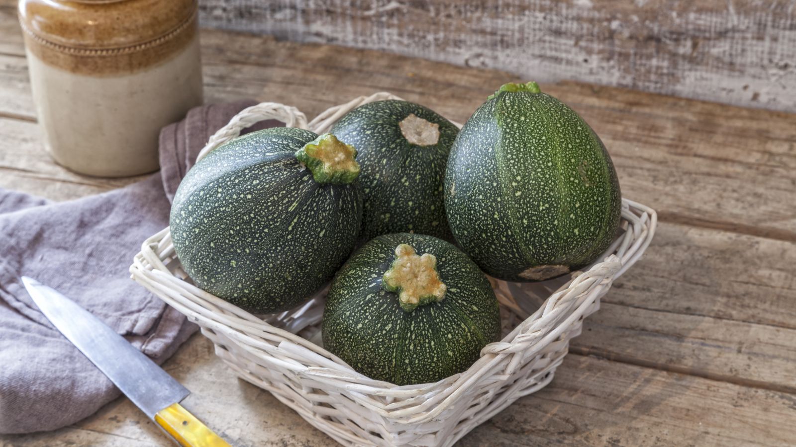 A close-up shot of a square woven basket filled with a small pile of round fruits of the Cucurbita pepo ‘Tondo di Piacenza’ variety, all placed on a wooden surface indoors