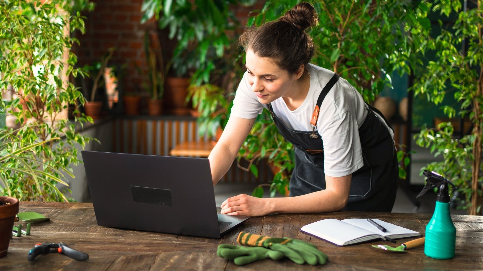 Young female gardener in black apron using a laptop on a wooden table surrounded by various potted plants in a cozy indoor room.