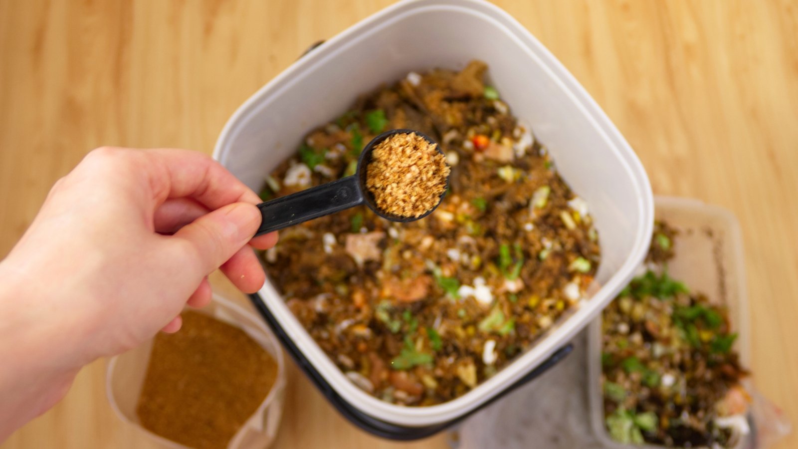A woman pours Bokashi Bran into a compost bin with kitchen waste using a black plastic spoon.