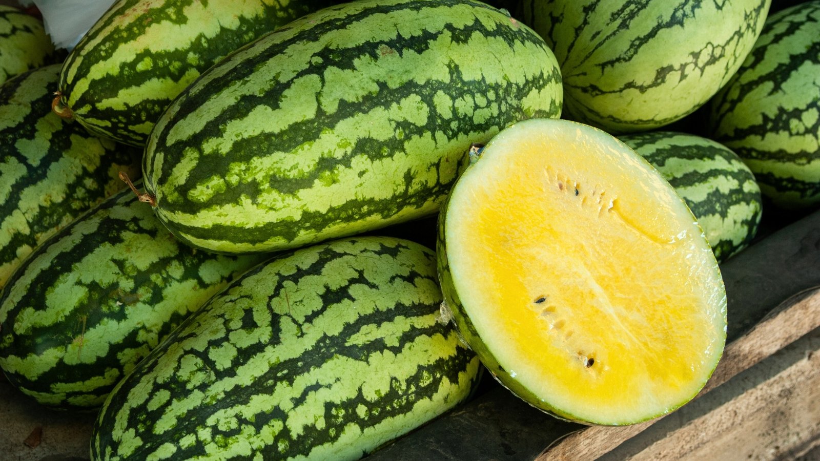 Harvested watermelons on a wooden table, oval with green rinds, one sliced open revealing bright yellow flesh.