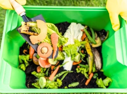 A gardener in yellow gloves puts kitchen scraps into a green compost bin full of soil and food scraps using a garden trowel, showing how composting can be done in a small space.