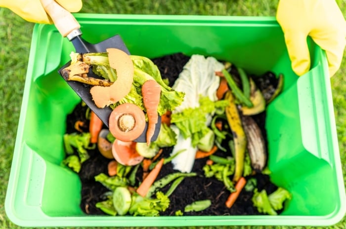 A gardener in yellow gloves puts kitchen scraps into a green compost bin full of soil and food scraps using a garden trowel, showing how composting can be done in a small space.
