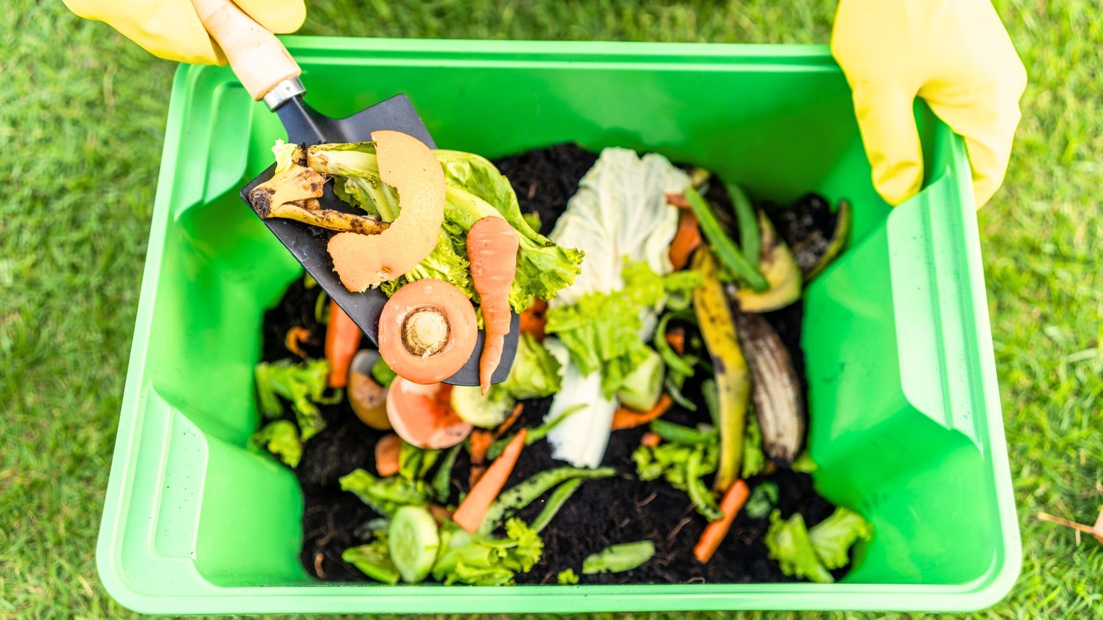 A gardener in yellow gloves puts kitchen scraps into a green compost bin full of soil and food scraps using a garden trowel, showing how composting can be done in a small space.