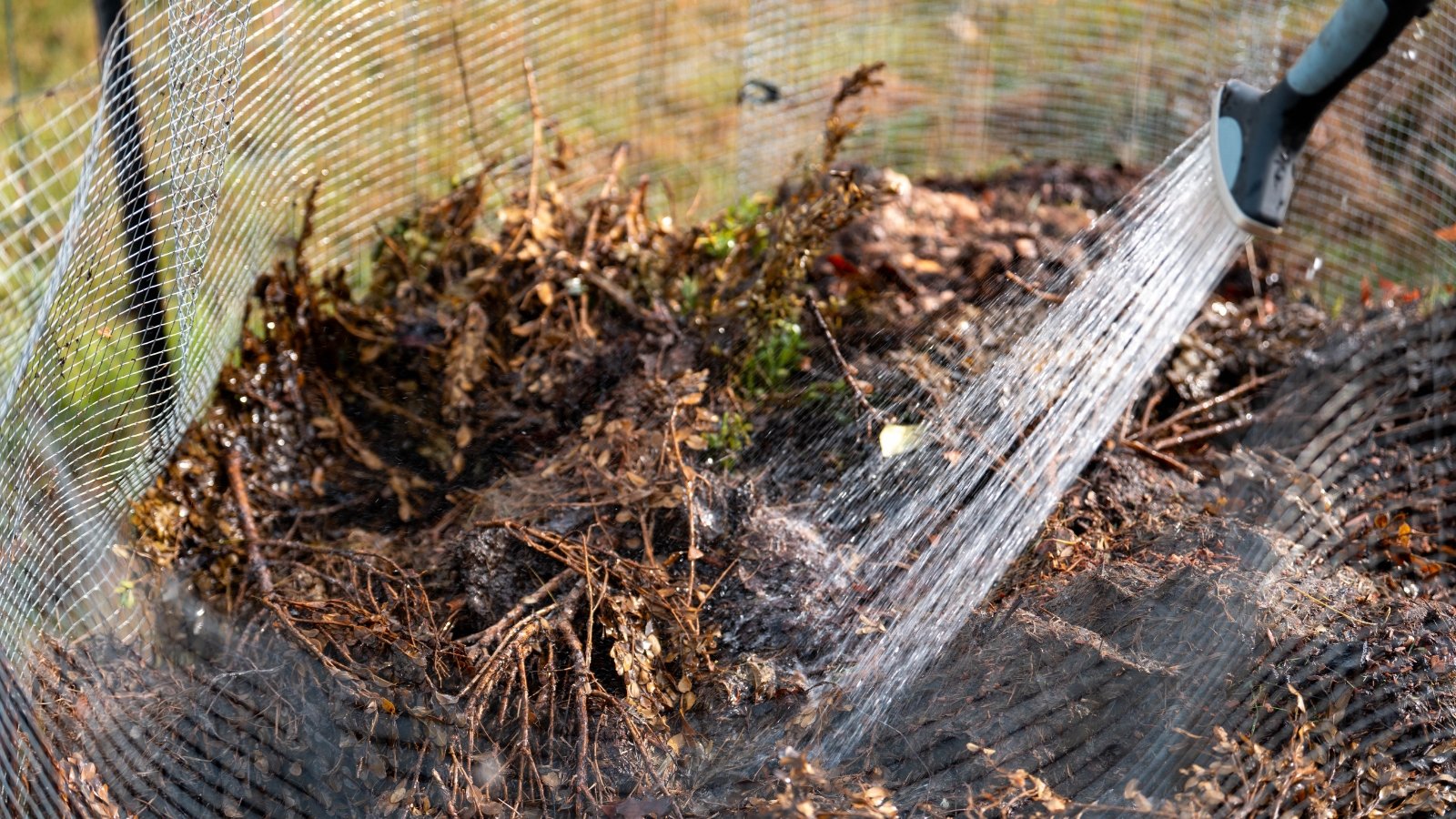 Close-up of a hose with a spray nozzle watering a compost pile enclosed with fine mesh.