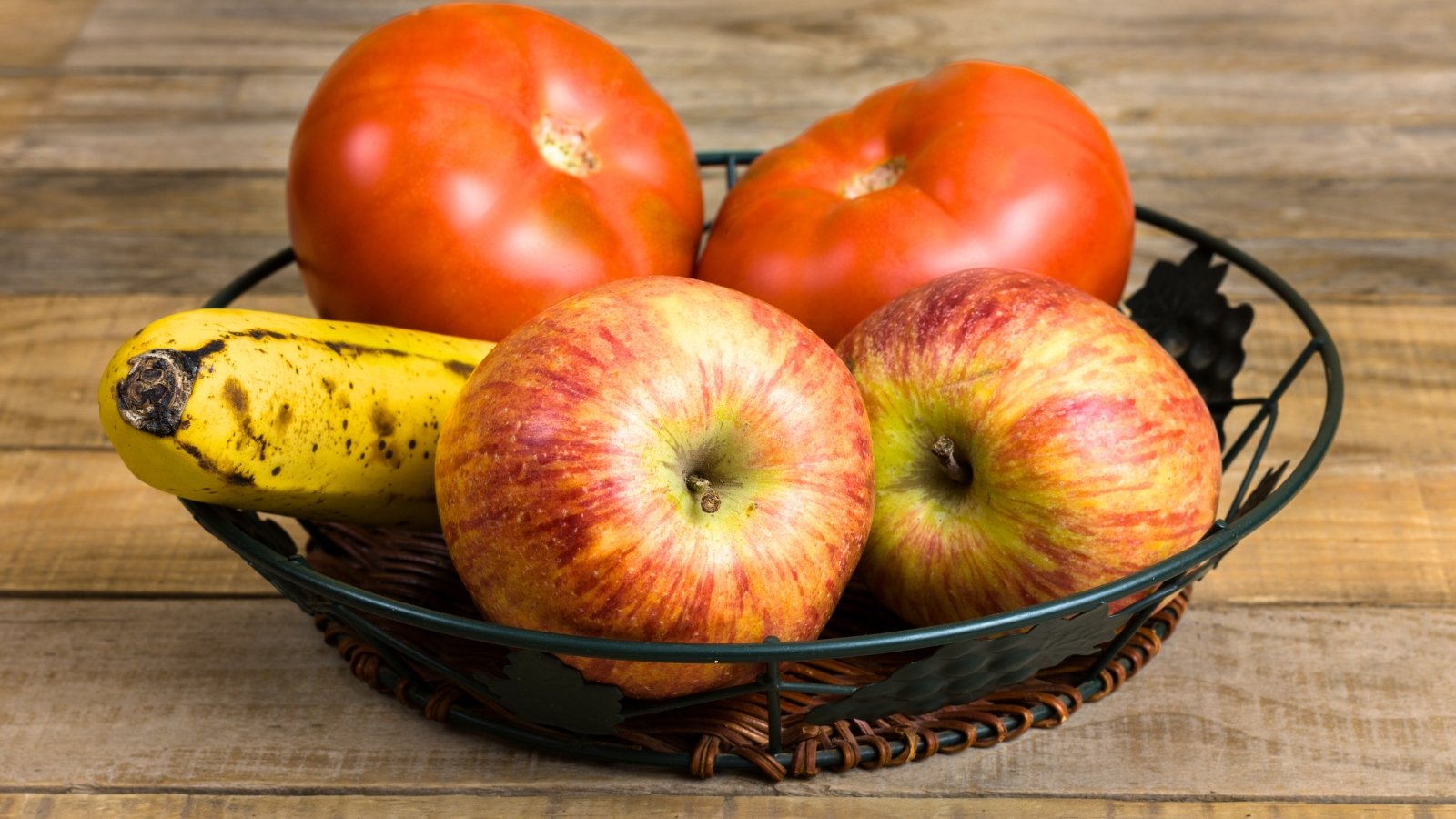 Close-up of ripe red-green apples, red tomatoes, and ripe bananas on a black decorative plate on a table.