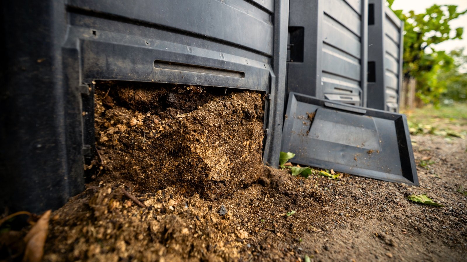 A row of black compost bins with the bottom lids open, spilling fresh brown compost.