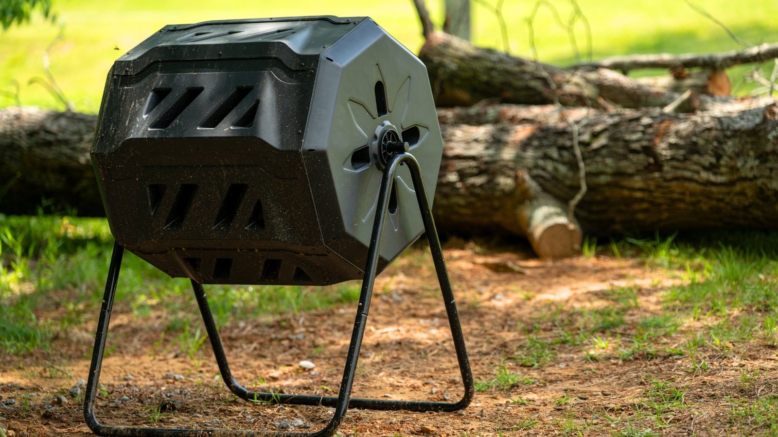 A black dual-chamber rotating compost tumbler on a stand surrounded by natural backyard greenery.