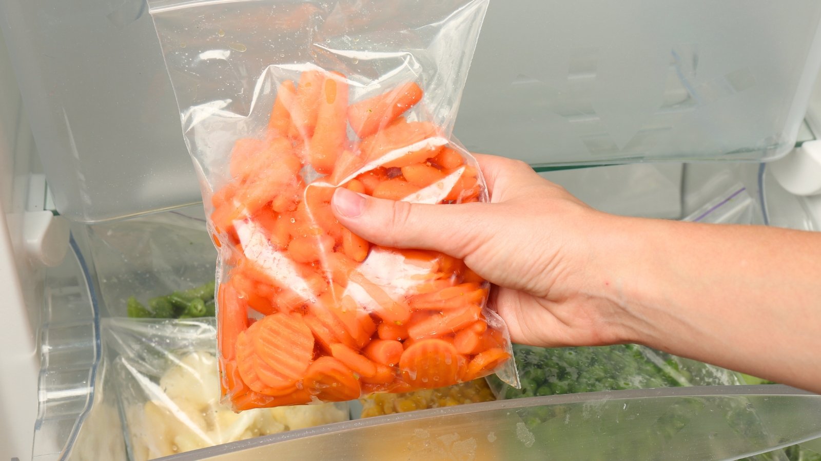 A woman's hand shows a plastic ziplock bag full of freshly frozen orange carrot pieces above a freezer.
