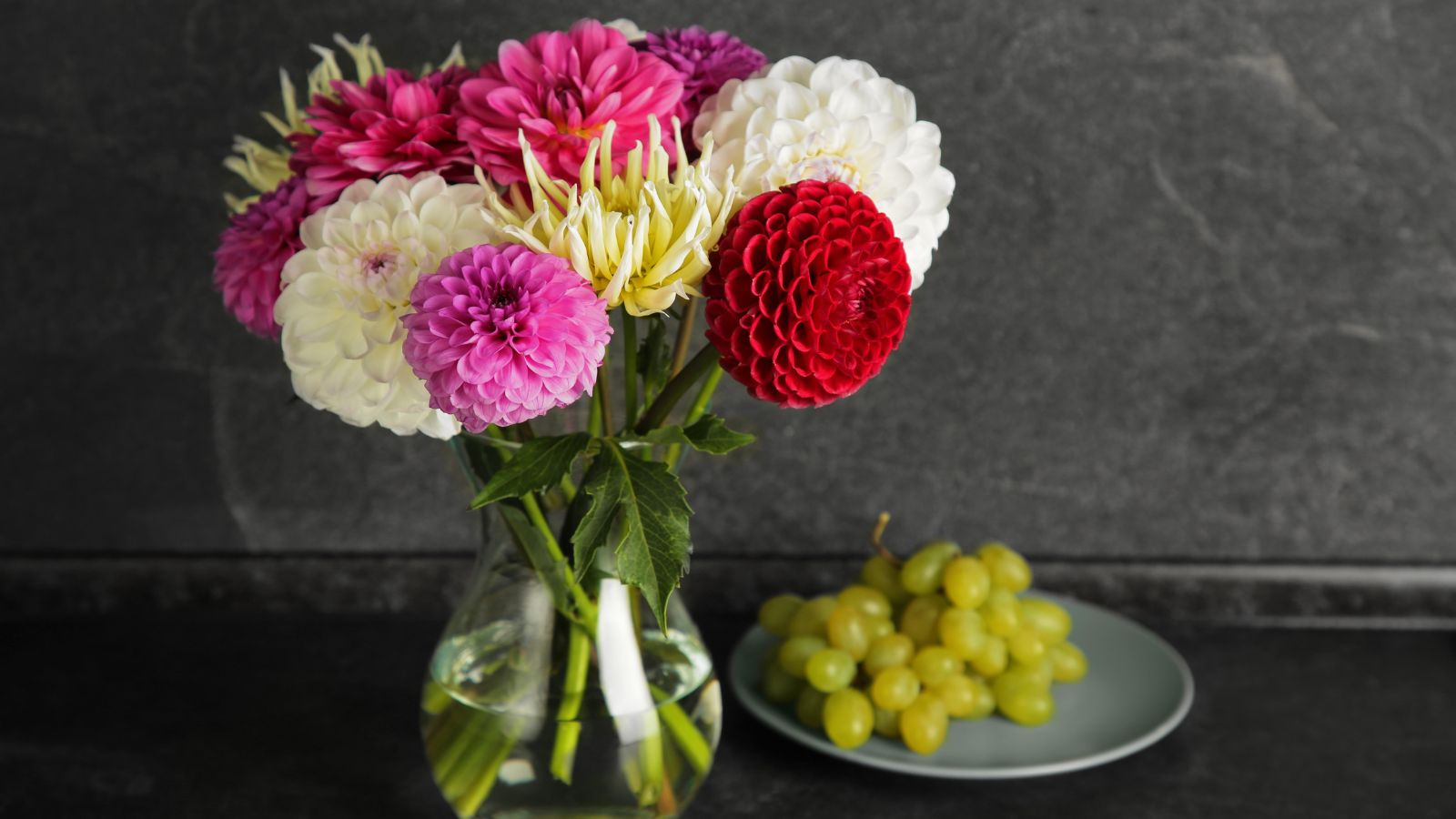 A close-up shot of a glass container with water and cuttings of several vibrant blooms in a well lit area indoors