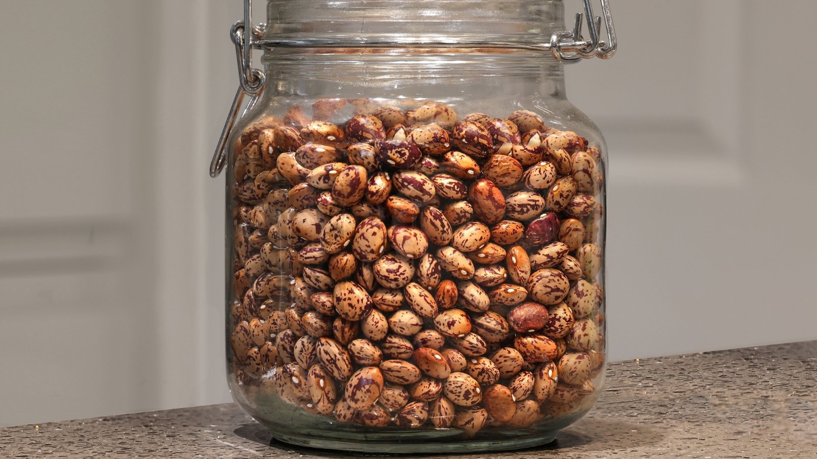 Borlotti beans with creamy skins and reddish streaks fill a clear glass jar, showcasing their speckled pattern.

