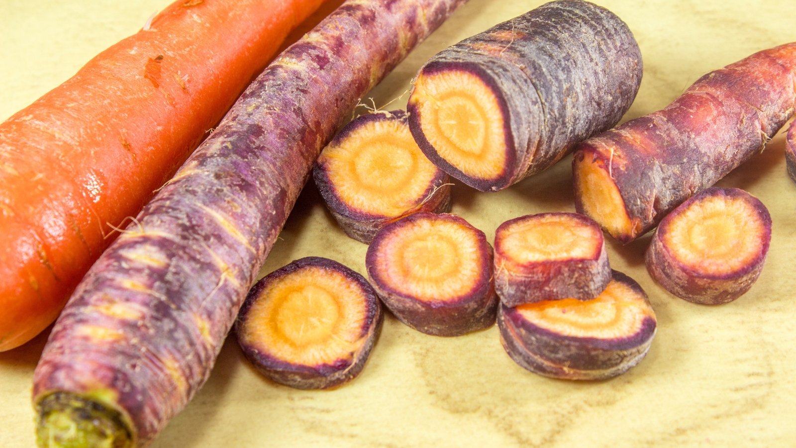 Cosmic Purple carrots with deep purple skins and vibrant orange centers are displayed on a wooden surface, with one sliced into round pieces showing the colorful contrast.