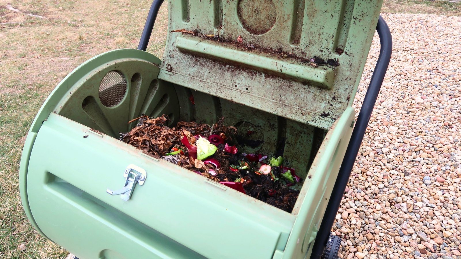 A green tumbler compost bin filled with a mix of colorful kitchen scraps like vegetable peels, fruit rinds, and wilted greens, visible through the open lid.