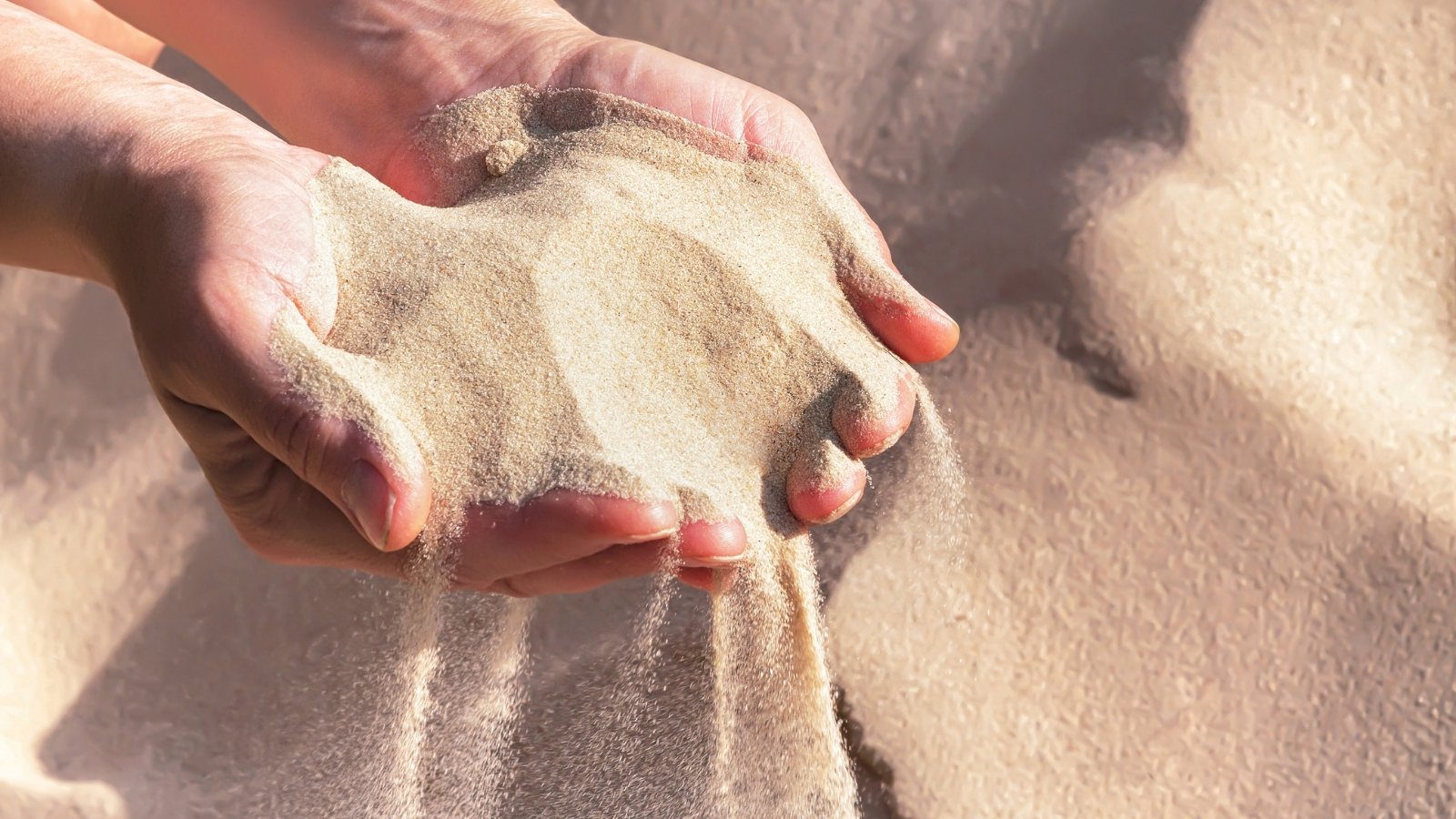 Close-up of pale beige sand flowing through a woman’s cupped hands against a soft, natural background.
