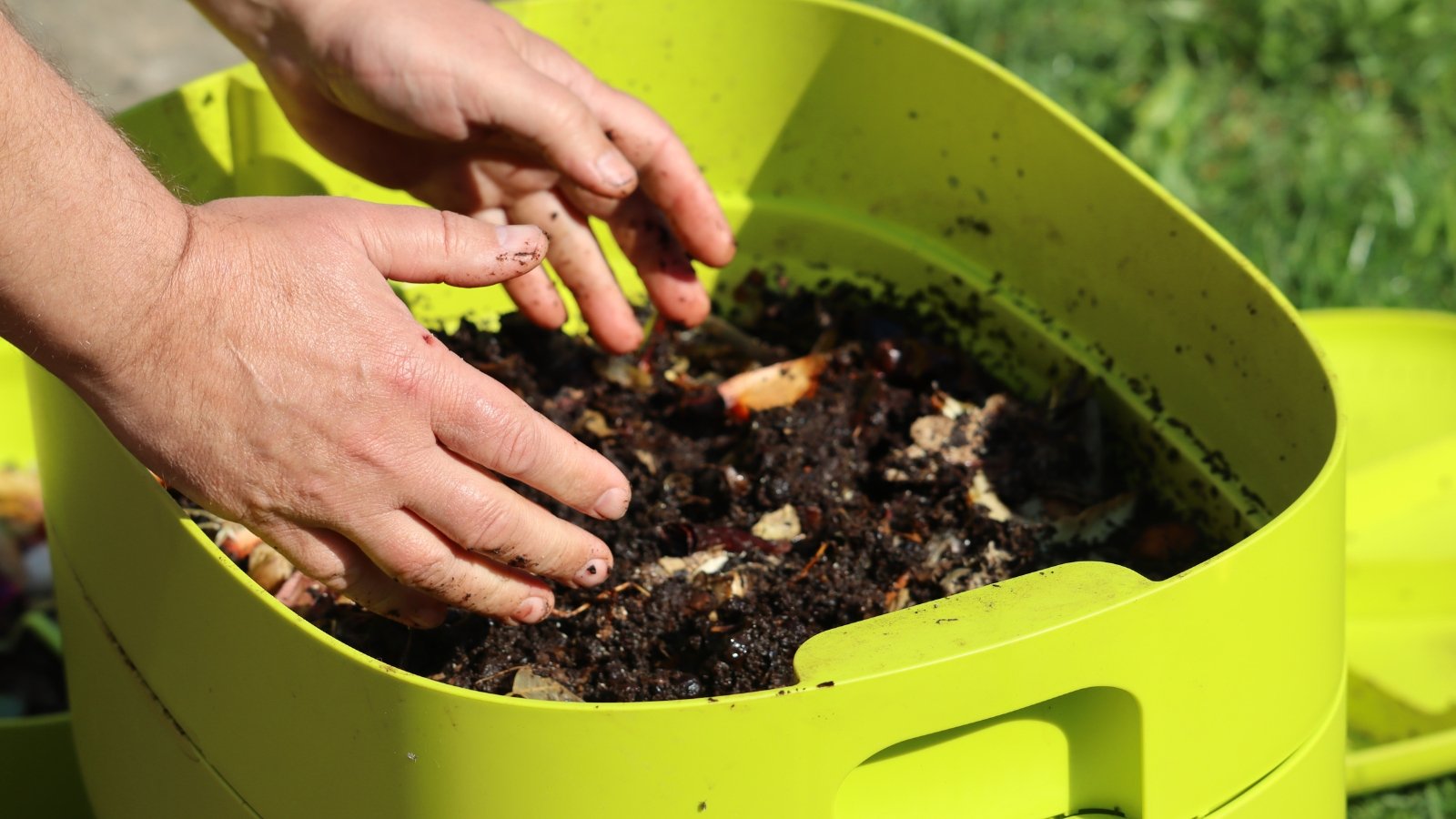 Close up of male hands tending to compost in a plastic green vermicomposter filled with black wet soil, food scraps and worms.