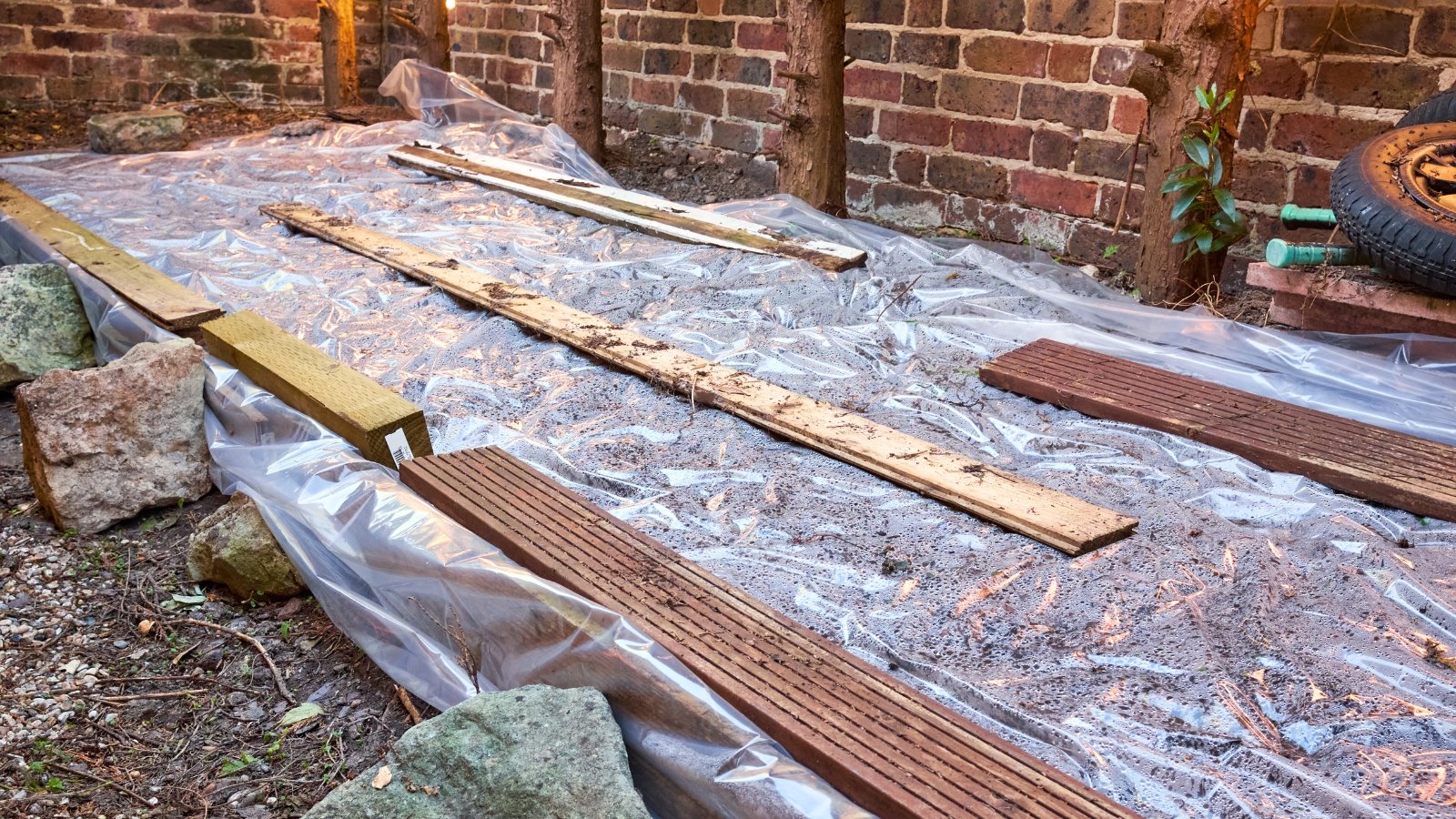 Transparent plastic fabric tightly covers garden soil, held down by wooden boards and large boulders near a wooden wall to solarize and kill weeds.
