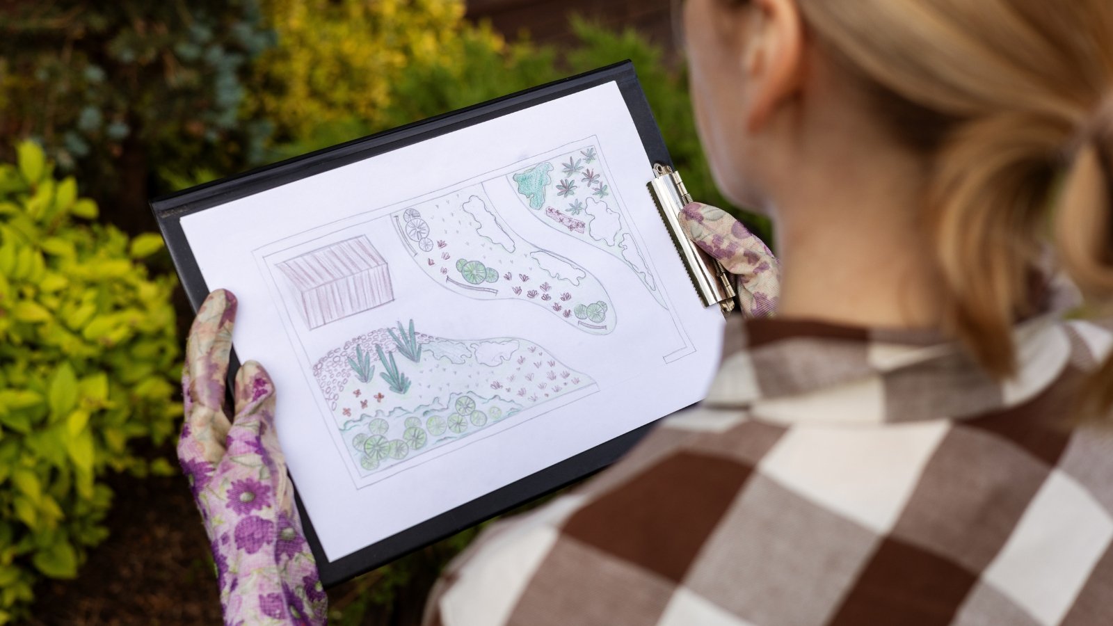 Close-up of a woman in a plaid shirt and flowered gloves holding a backyard map.
