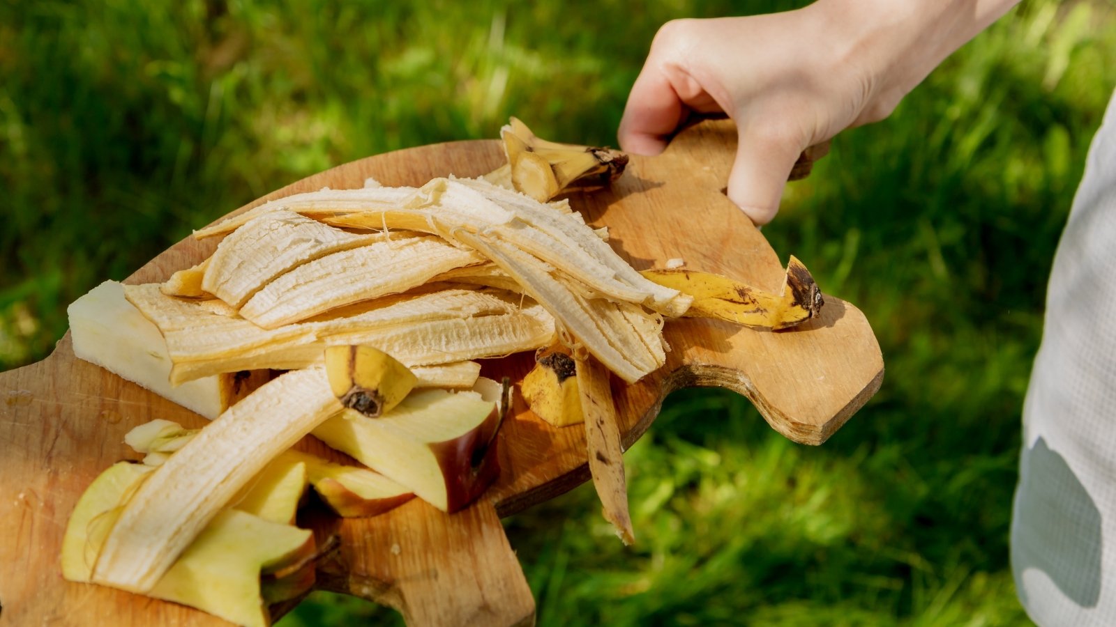 A woman holds a wooden cutting board full of banana peels and apple pieces.