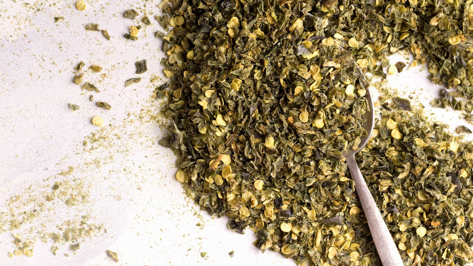 Tiny dry green jalapeno pepper pieces rest in a white surface and a spoon, showing a coarse, crumbly texture.
