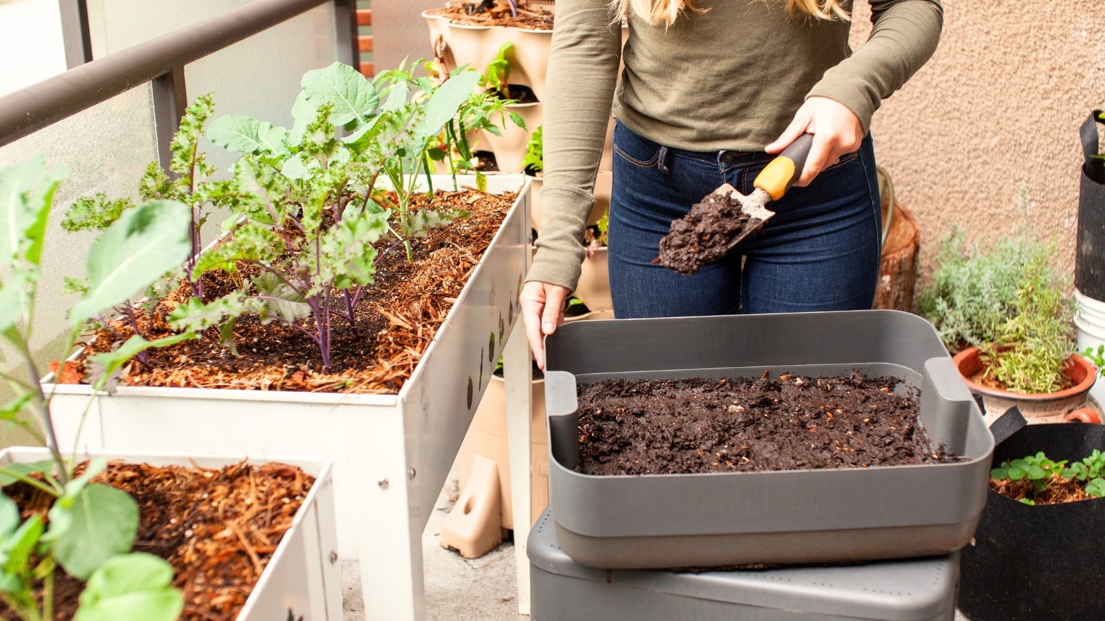 A woman stands by a compost bin with a small garden trowel full of vermicompost, adding it to young plants growing in raised beds on a balcony.