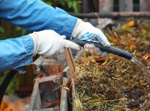 A male gardener in white gloves and a blue denim shirt waters a large compost heap with a black hose to make it wet in the garden.