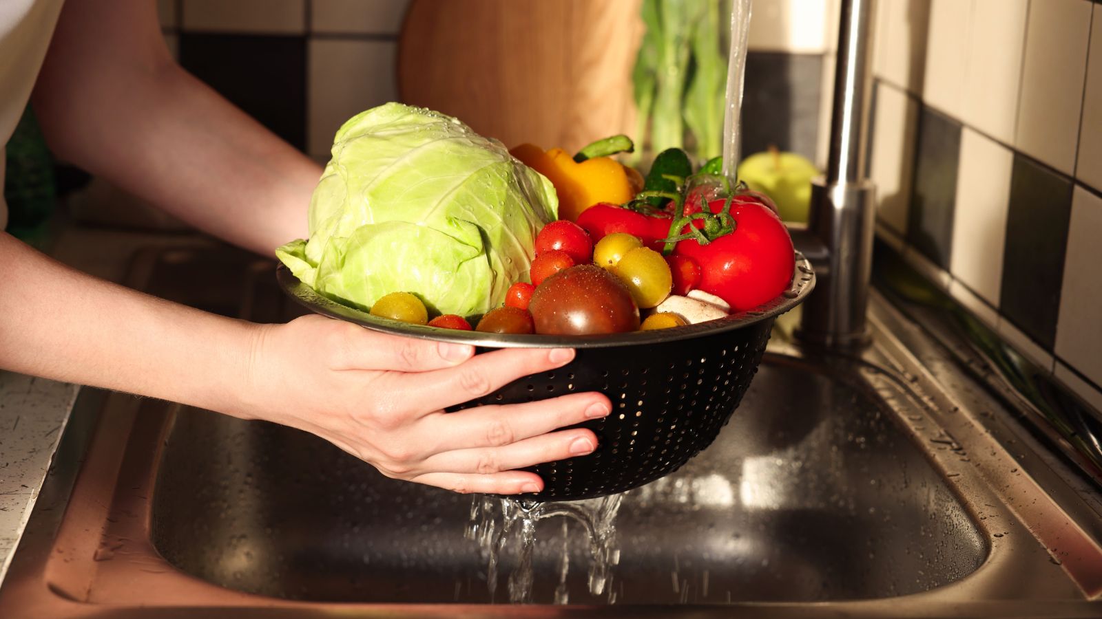 A close-up shot of a person holding a colander filled with fruits and vegetables, in the process of washing them over a sink