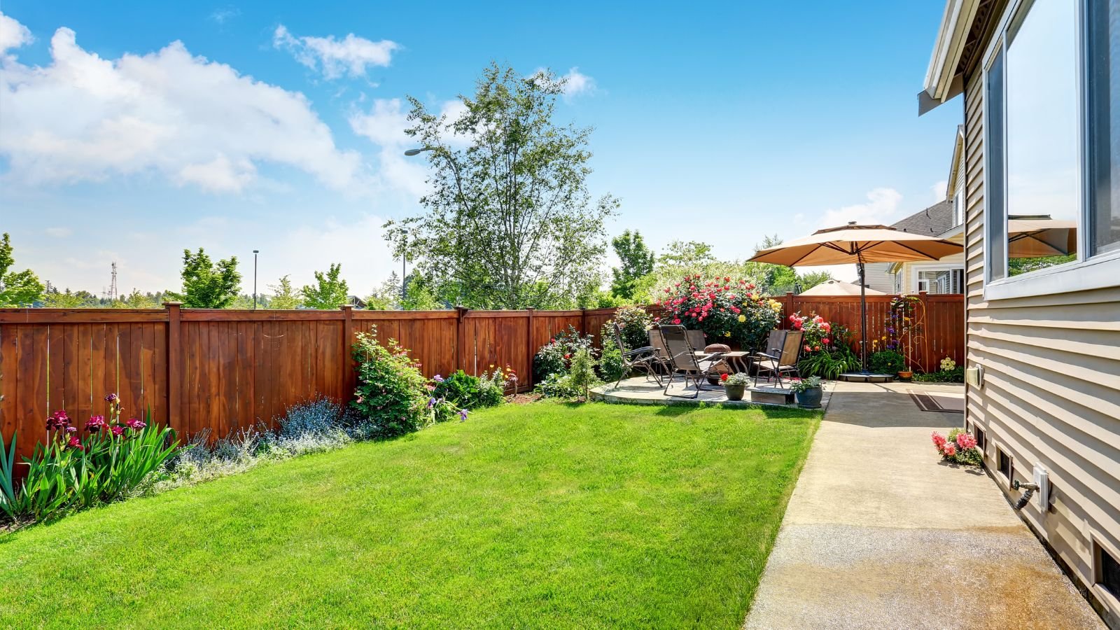 A shot of a large, landscaped backyard area near a home, showcasing various flowers and plants, alongside patio chairs beside a pathway near the house, all situated in a well lit area outdoors