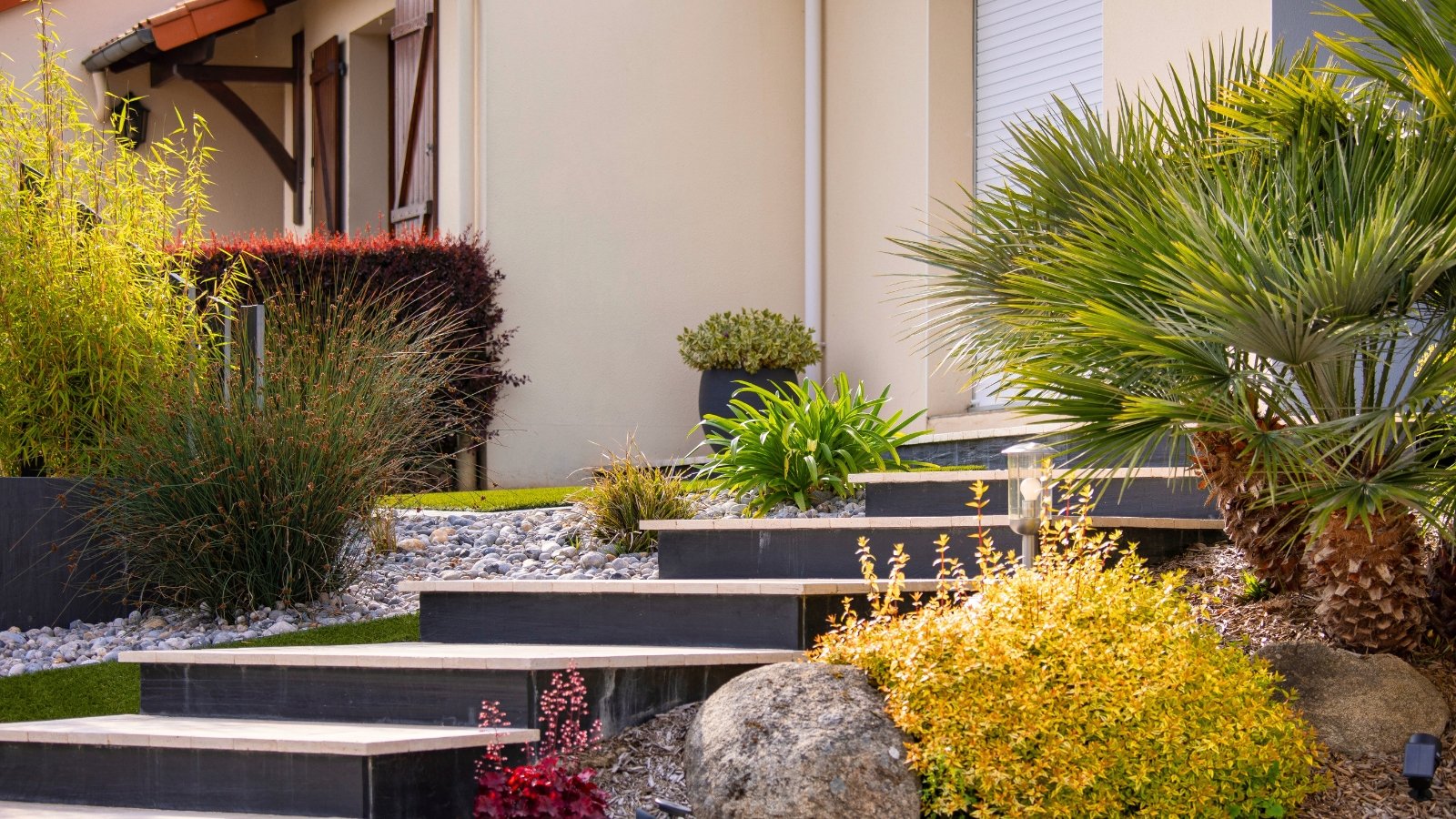 Charming small landscaped garden featuring Juncus patens, low palm, flowering Heuchera, climbing Ligustrum sinense, and other plants on a bed of small gray pebbles.