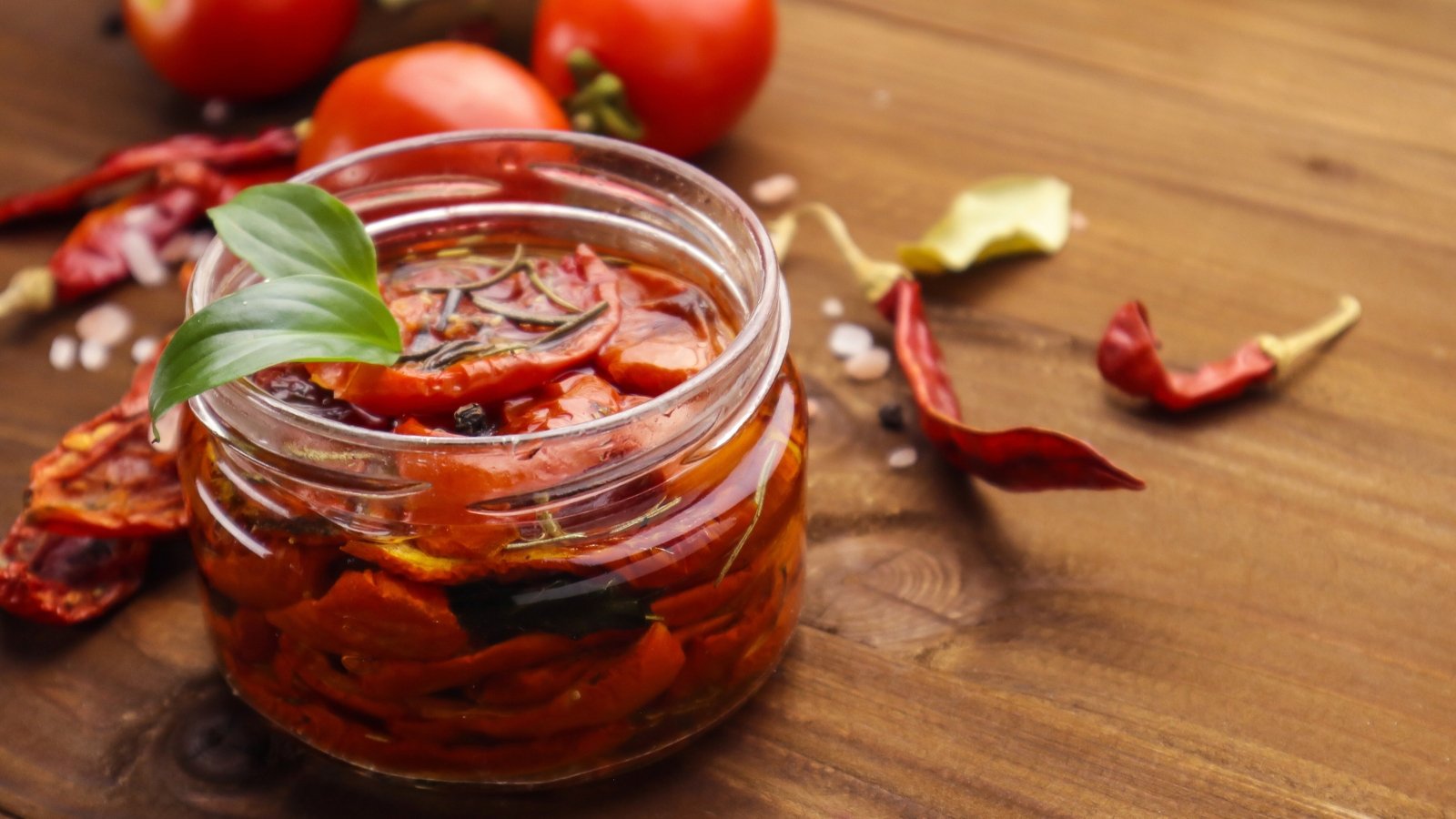 Glass jar filled with sun-dried tomatoes submerged in golden olive oil, placed on a wooden table.