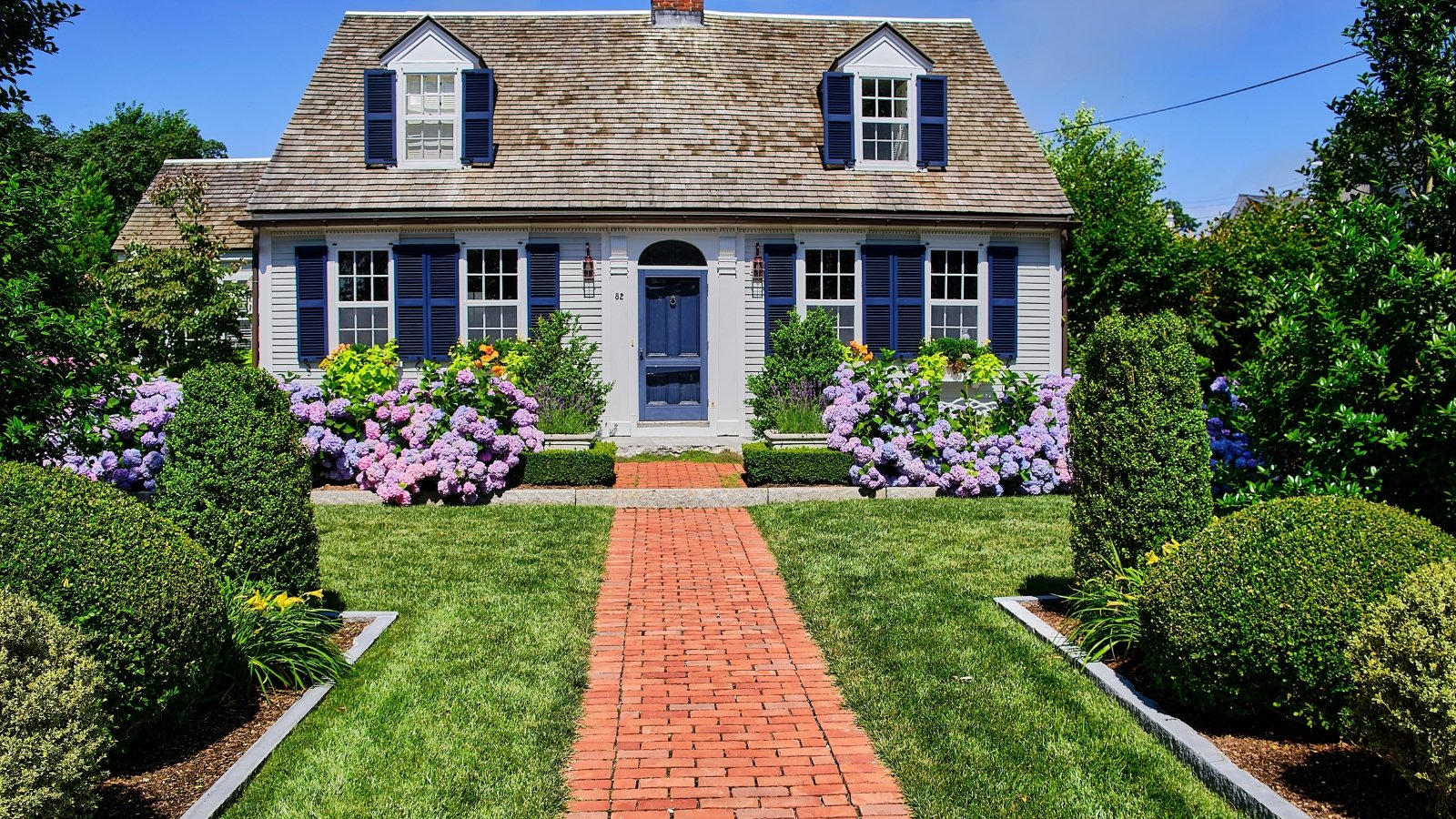A large, elegant white house with a gray-brown roof and blue shutters and doors is surrounded by flower beds filled with neatly trimmed boxwood and lush blooming hydrangea bushes, set against a manicured green lawn divided by an orange stone path.