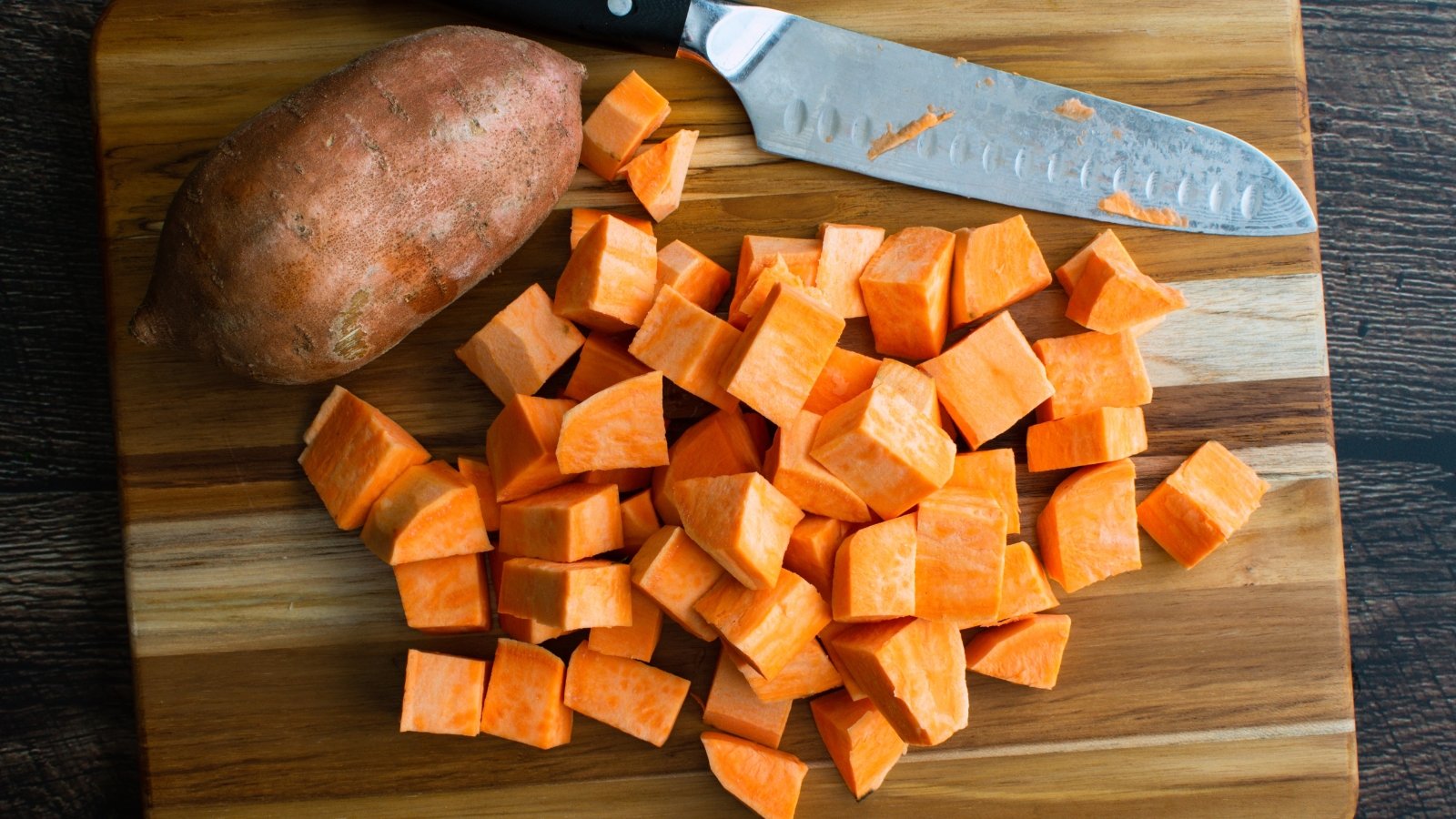 Large orange-fleshed sweet potato cubes rest on a wooden board beside a large kitchen knife, ready for freezing.