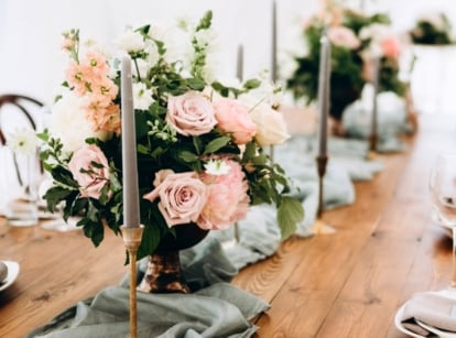 Wedding table decorated with gold candlesticks and elegant floral arrangements of pastel pink roses, pale peach delphiniums, pink-beige dahlias, and white flowers arranged in gold vases, showcasing our flower selection for Taylor Swift’s wedding.