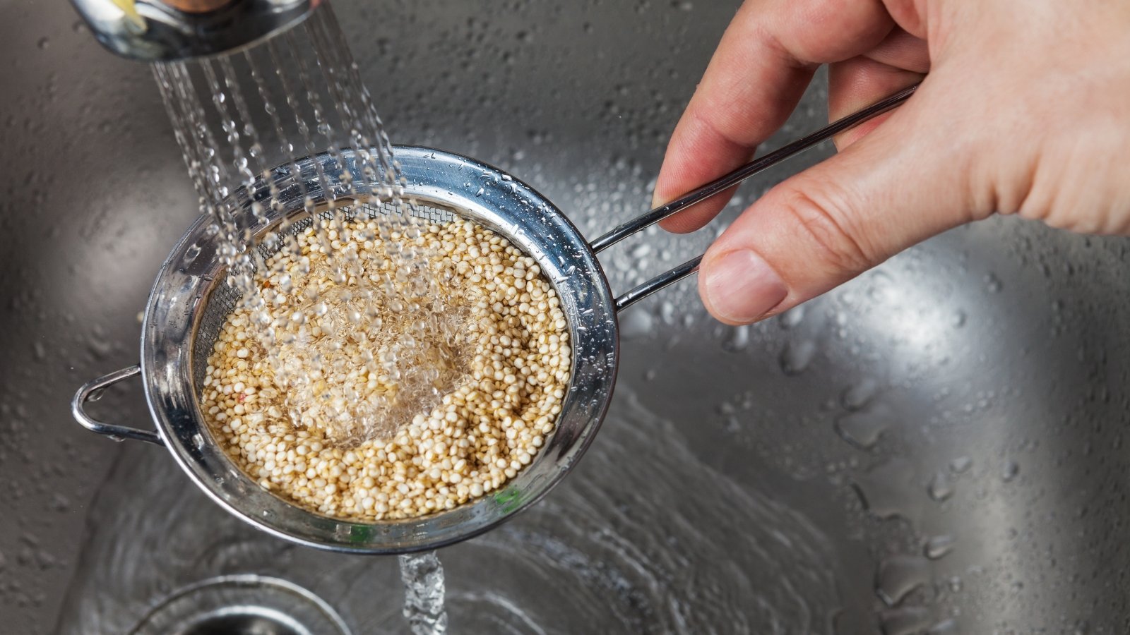 A woman’s hands wash tiny, round beige-white seeds in a small colander under running tap water in the sink.