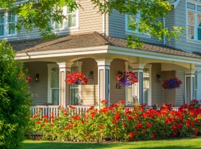 A gorgeous large soft blue house is surrounded by a green lawn, brightly blooming flower beds, tall trees, and bushes, with its porch decorated by hanging baskets of red and purple flowers, showcasing easy landscaping tweaks that enhance curb appeal.