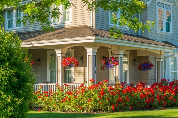 A gorgeous large soft blue house is surrounded by a green lawn, brightly blooming flower beds, tall trees, and bushes, with its porch decorated by hanging baskets of red and purple flowers, showcasing easy landscaping tweaks that enhance curb appeal.